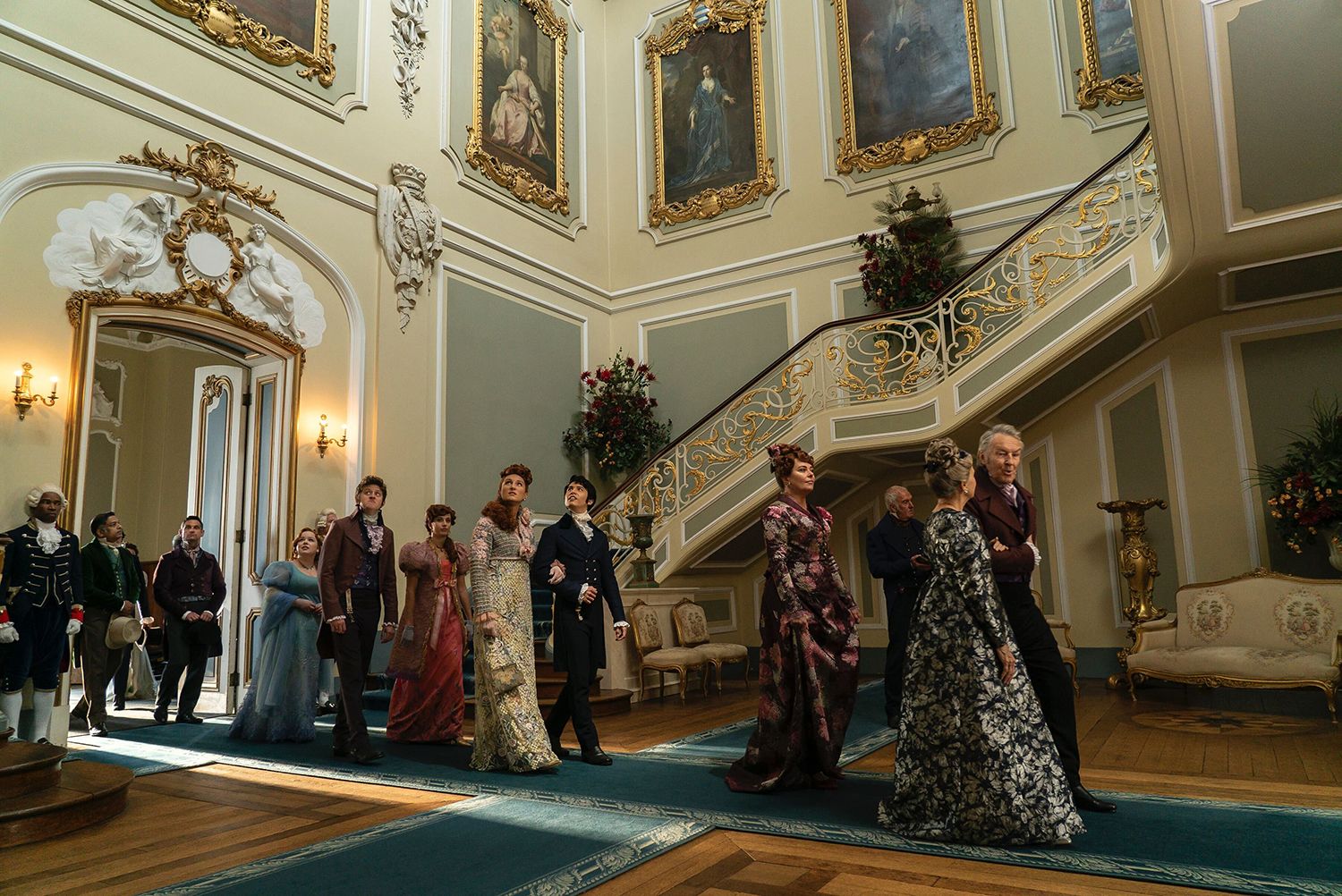The characters in Bridgerton walk through the imposing hall, past a grand staircase at Wrest Park. Several of them are looking up at the impressive paintings that line the walls.