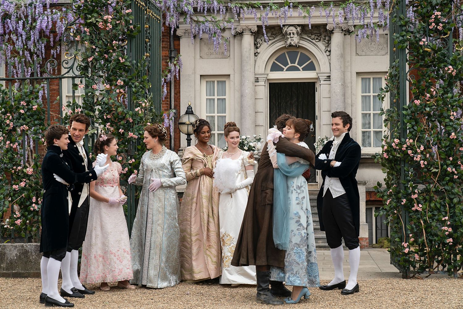 The Bridgerton family are standing outside their home, wearing smart evening wear. Colin and Eloise embrace to one side, while other members of the family look on and smile.