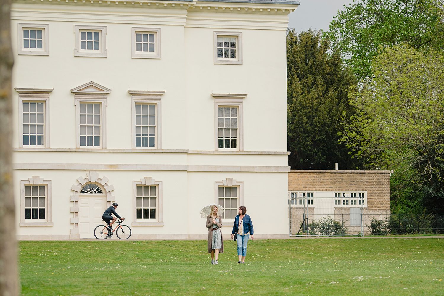 Two women in modern dress walk in the grounds of Marble Hill, one carrying an umbrella. They are watched by a cyclist, who is riding in front of the house.