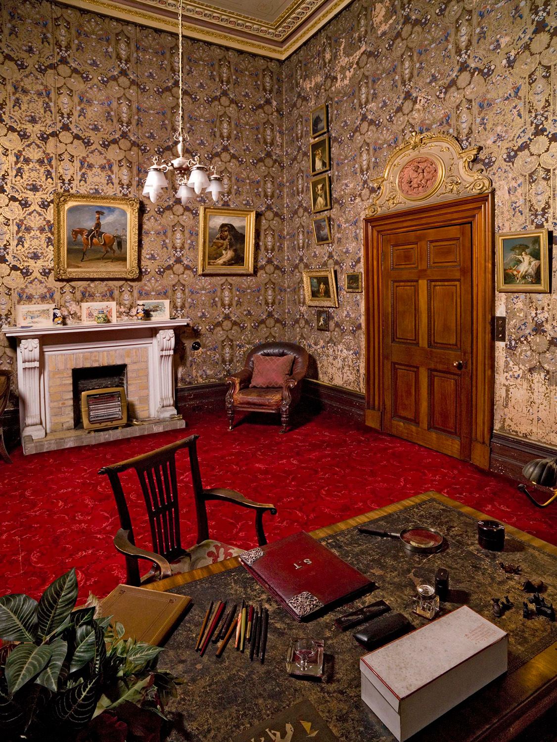 The morning room at Brodsworth with a deep red carpet and a comfy-looking brown leather armchair next to a white mantelpiece above a fireplace. There are animal portraits on the walls behind the chair.