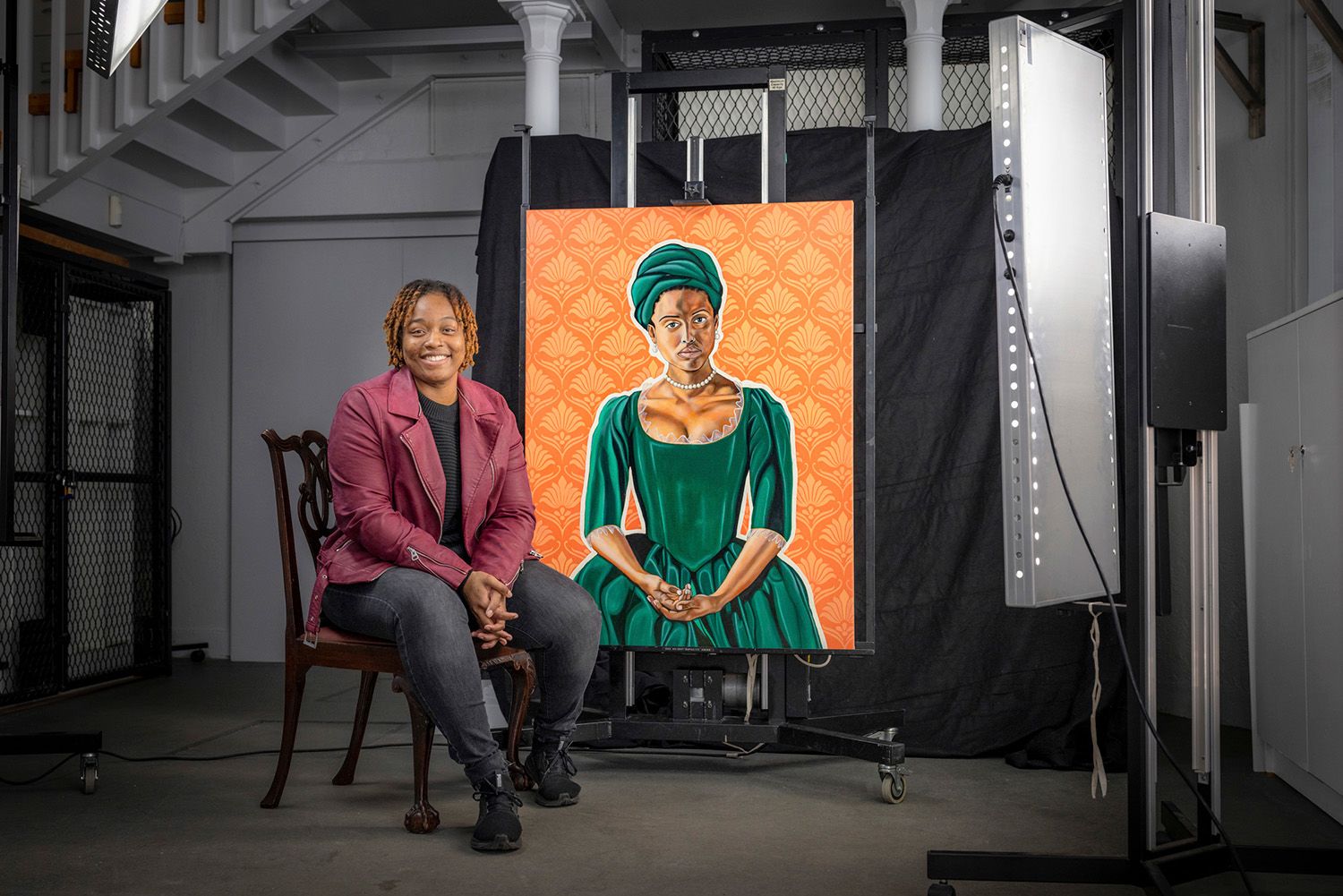 The artist Mikéla Henry-Lowe wearing a maroon leather jacket and black jeans sits smiling broadly next to her painting of Dido Belle.