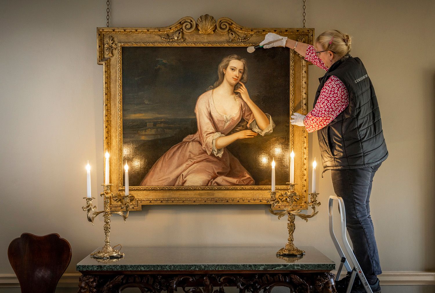 A conservator wearing white gloves and a gilet, cleans a portrait of Henrietta Howard with a delicate brush. She's standing on a ladder above three lit candles at Marble Hill.