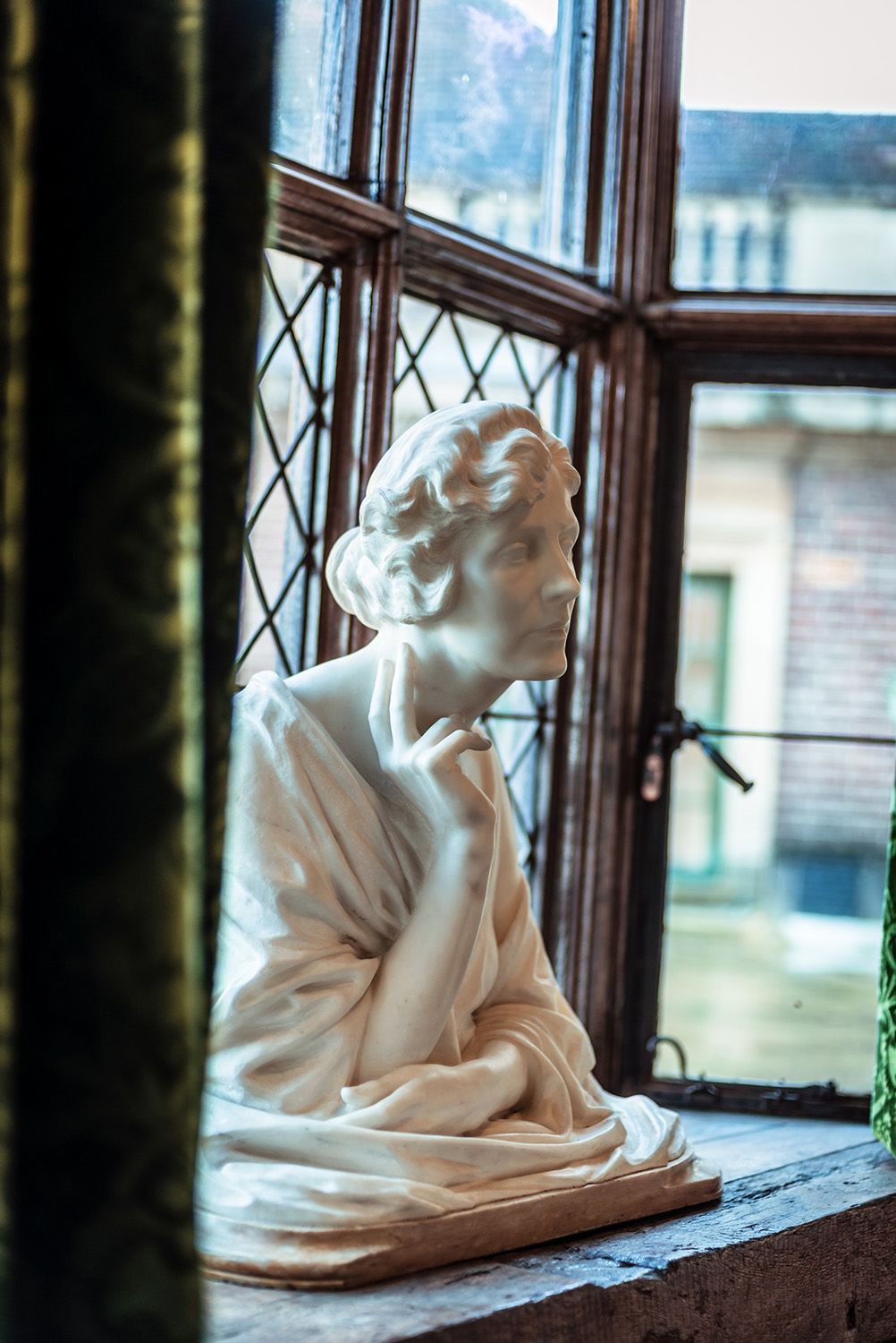 A side view of the bust of Virginia Coutauld, showing the two fingers raised to the side of her throat in a latticed window at Eltham Palace.