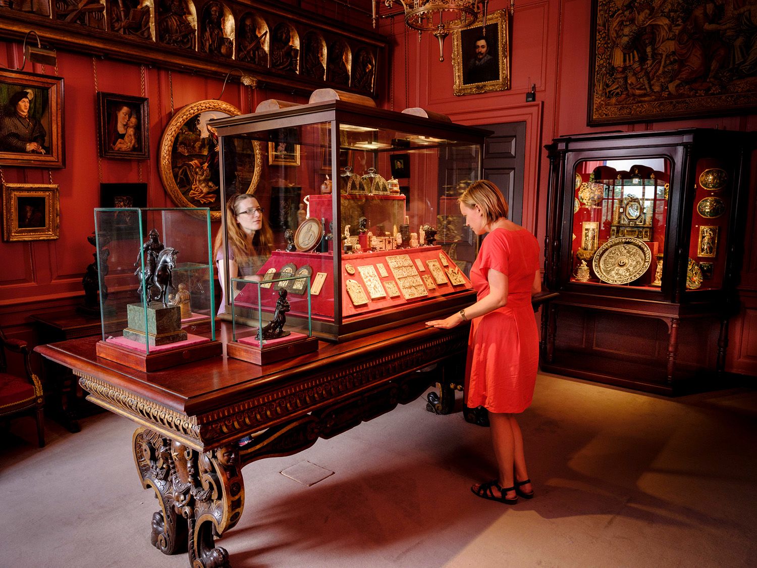 Two women look at objects from The Wernher Collection in glass cases in a dark red room in Ranger's House.