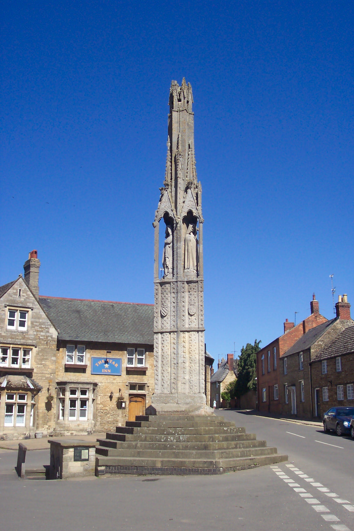A photo of a tall, stone monument with two Queen Eleanor figures visible, in a bright blue Northampton sky.