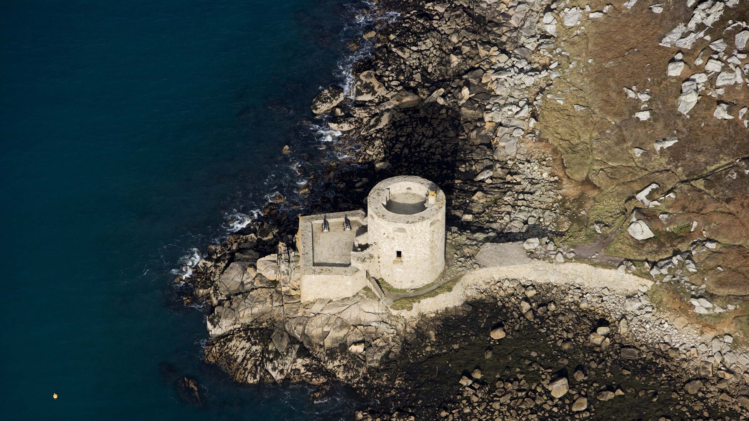 Aerial photograph of a small stone tower and outbuilding perched on a rocky coastline